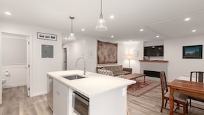 Une cuisine moderne avec un îlot central, un coin repas avec une table et des chaises en bois, un salon avec un canapé et une cheminée, et un téléviseur fixé au mur.
