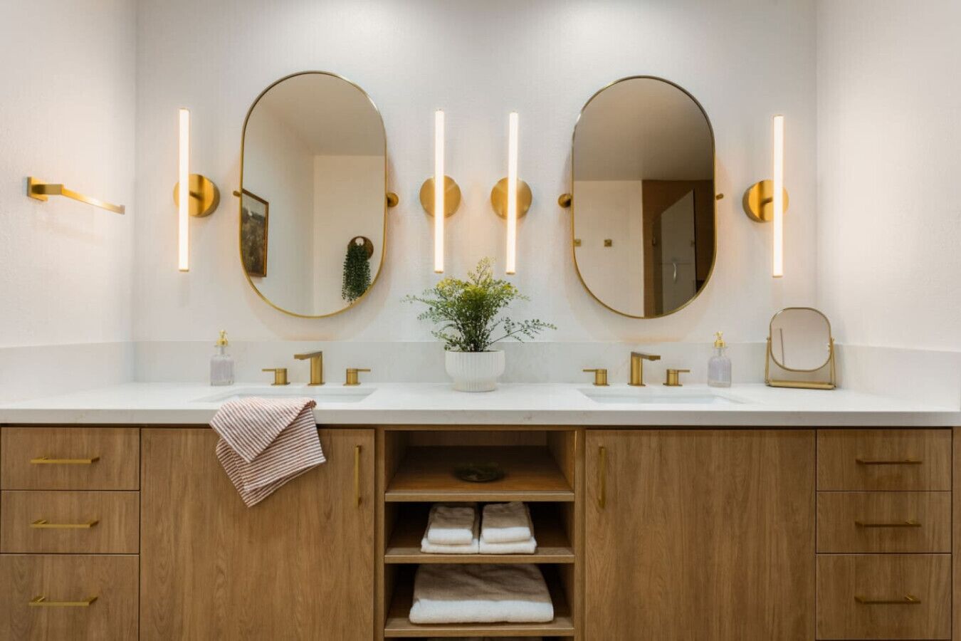 A bathroom with two sinks, gold fixtures, and a mirror. There are two wall-mounted lights and a plant on the counter.