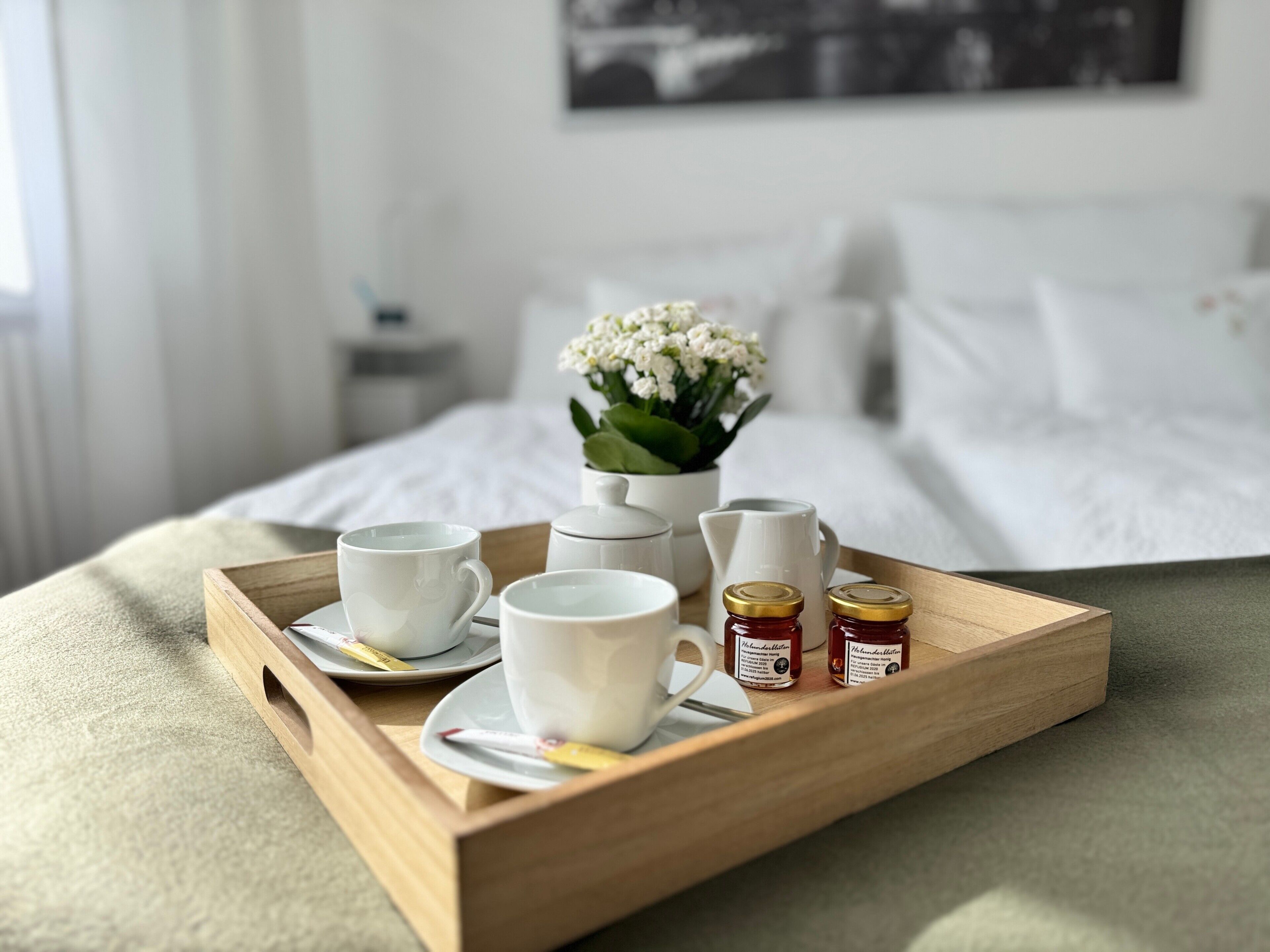 Uma bandeja de madeira com xícaras e potes de mel sobre uma cama com roupa de cama branca e um vaso de flores branco.