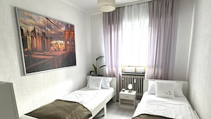 A hotel room with two beds, a radiator, a nightstand with a lamp, a window with curtains, and a cityscape painting on the wall. - Refugium 2020 - The vacation apartment - 69 sqm with balcony, perfect for 4 people, max. 7 (Marienheide)