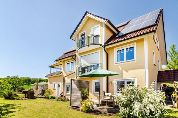 A multi-story house with a balcony, a patio with outdoor furniture, and a garden.