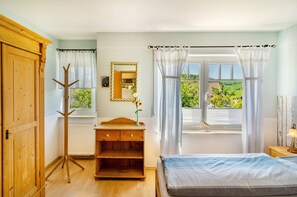 A bedroom with a wooden wardrobe, a bedside table with a lamp, a window with curtains, and a view of greenery outside.