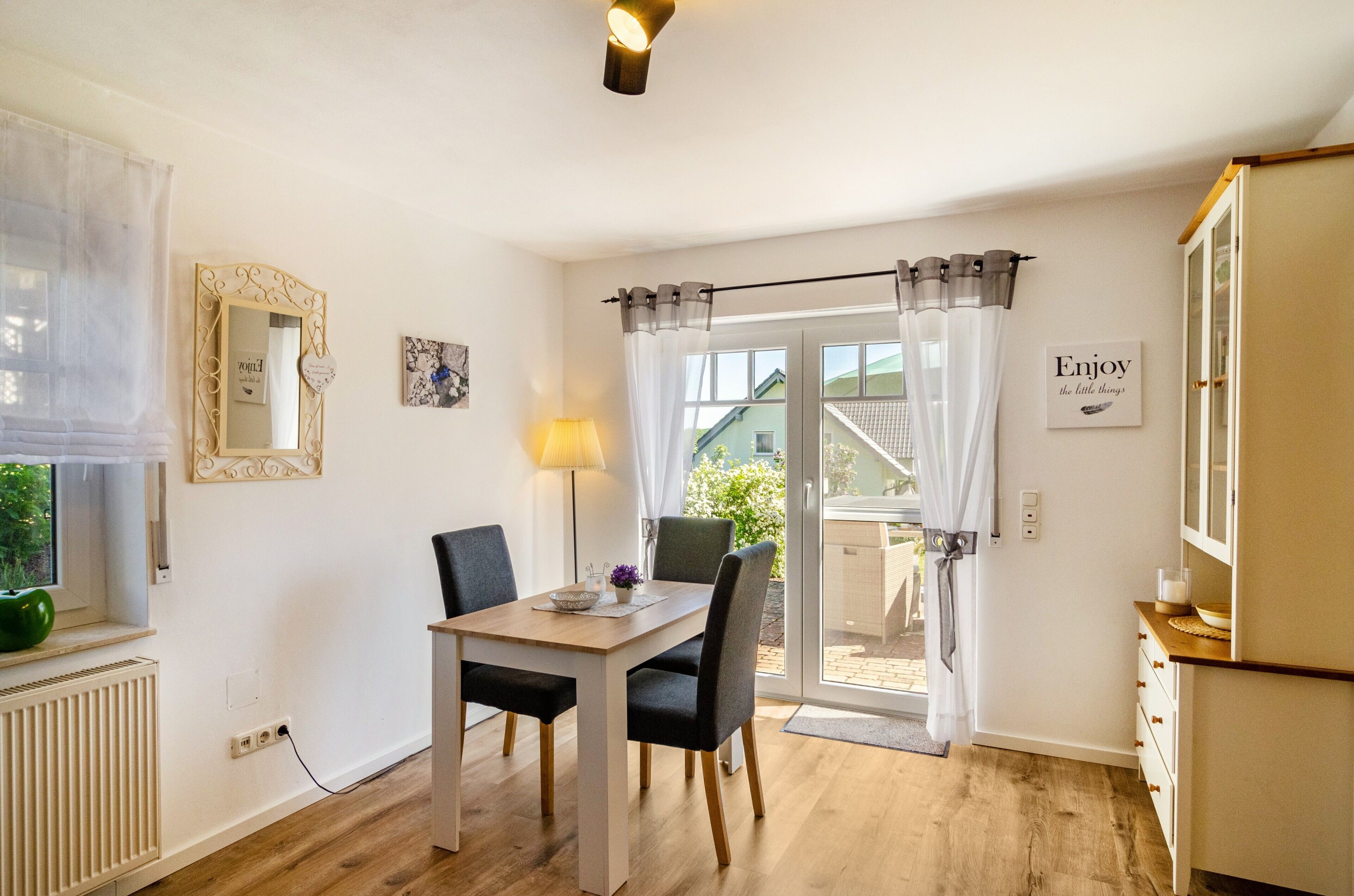 Ein Zimmer mit Esstisch, Stühlen, einer Lampe, einem Spiegel, einem Fenster mit Vorhängen, einer zur Außenwelt führenden Tür sowie einer Wand mit einem gerahmten Bild und einem Schild.