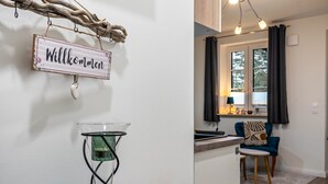 Un interno moderno con un cartello di benvenuto, un tavolo in vetro con un contenitore d'acqua, un tavolo da pranzo in legno, una sedia blu e vista su un angolo cottura.