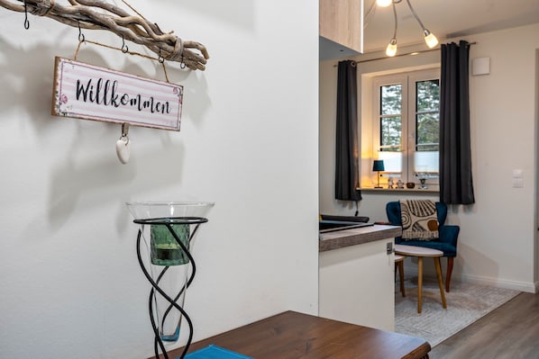A modern interior with a welcome sign, a glass table with a water container, a wooden dining table, a blue chair, and a view of a kitchen area.
