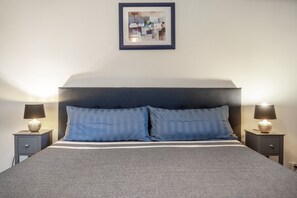 Una cama bien tendida con un cabecero oscuro, dos mesitas de noche con lámparas, y un cuadro enmarcado en la pared.