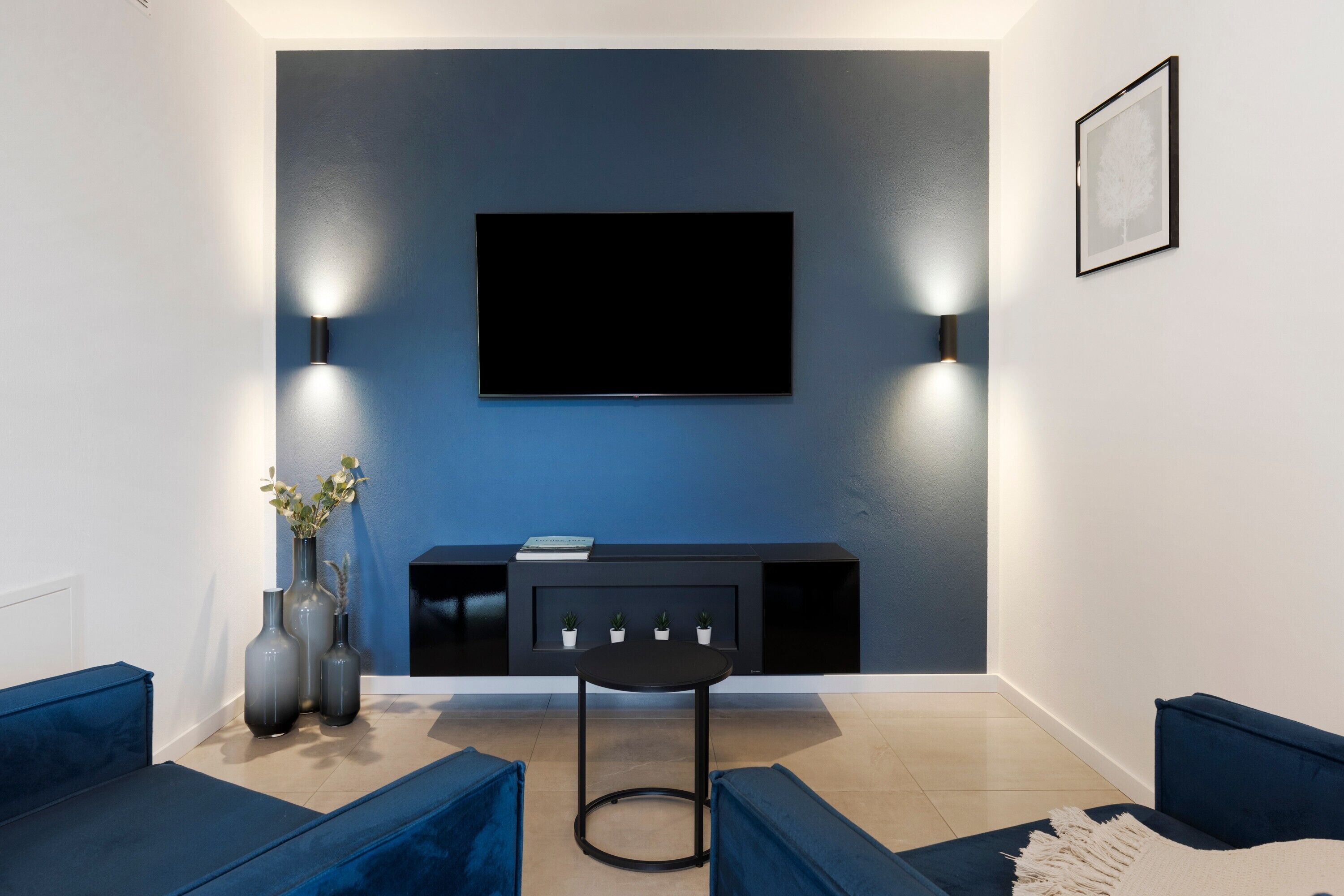 Ein modernes Wohnzimmer mit einem Flachbildfernseher an einer dunklen Wand, einer schwarzen Unterhaltungseinheit darunter und zwei Wandleuchten.