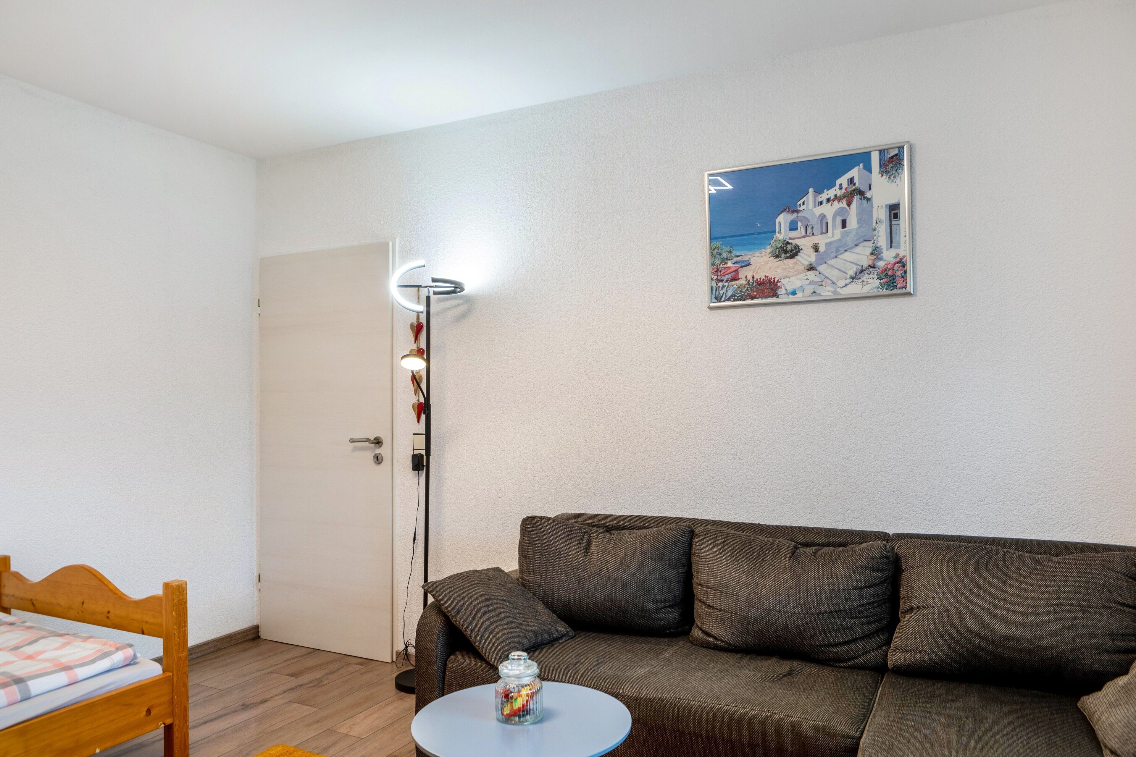 Una habitación con un sofá marrón, una mesita redonda pequeña, una silla de madera, una puerta blanca y un cuadro con una escena costera.
