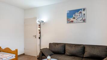 Una habitación con un sofá marrón, una mesita redonda pequeña, una silla de madera, una puerta blanca y un cuadro con una escena costera.