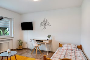 Quarto com cama, escrivaninha com cadeira, televisão na parede, janela e radiador.