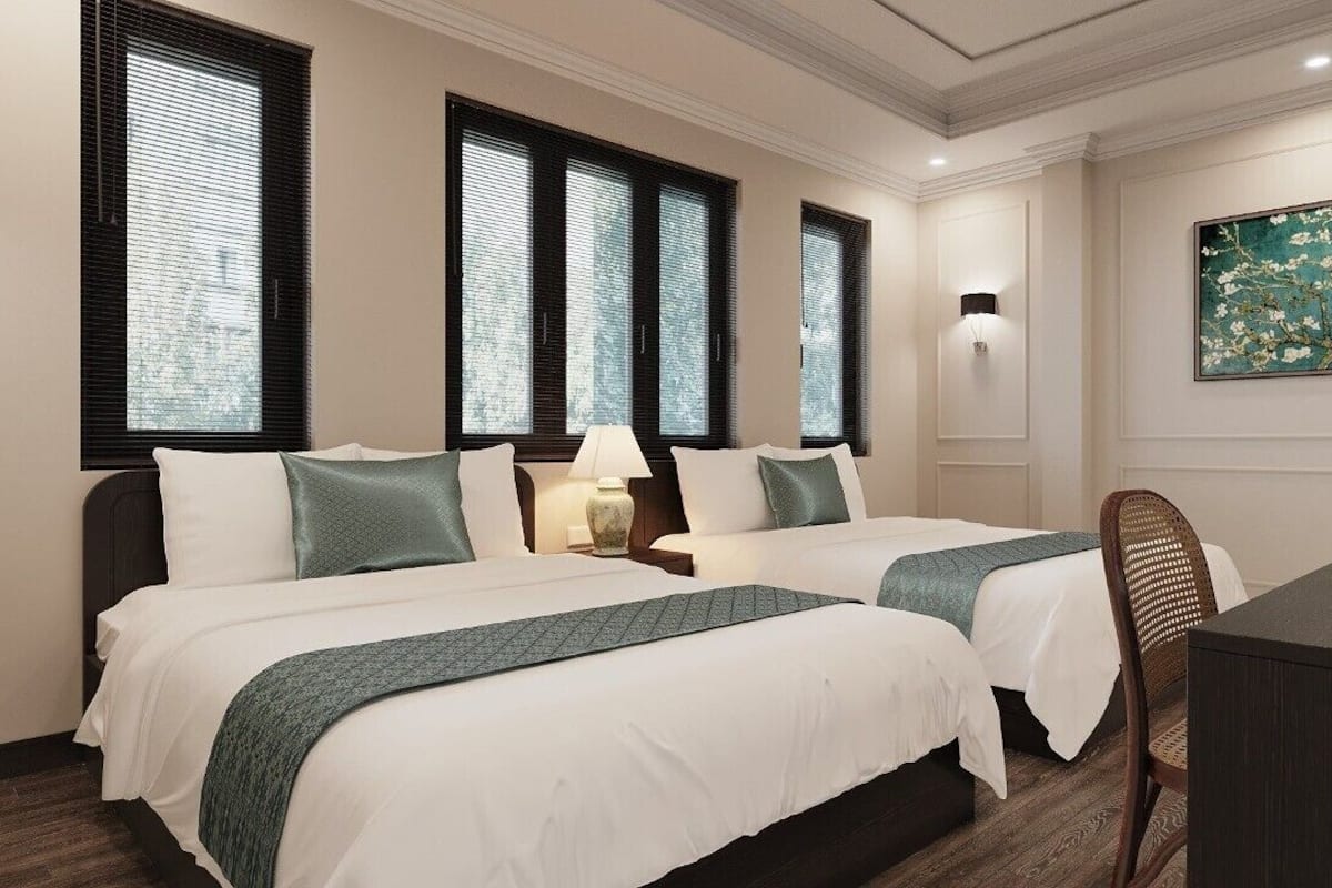 Ein Hotelzimmer mit zwei Betten, einem Schreibtisch und einem Stuhl. Drei Fenster mit Jalousien, ein Bild an der Wand und ein Blumenvasen auf dem Schreibtisch.