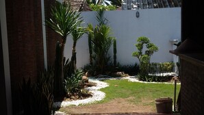 A covered patio area with a wooden floor, surrounded by a white wall and a brick wall. There are potted plants and a small garden area with a pathway. - Roots Hotel Trawangan (Gili Trawangan)