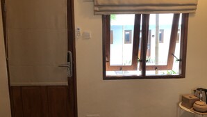 A room with a wooden door, a window with a view of the outside, a small table with a basket, and a wall-mounted thermostat. - Roots Hotel Trawangan (Gili Trawangan)