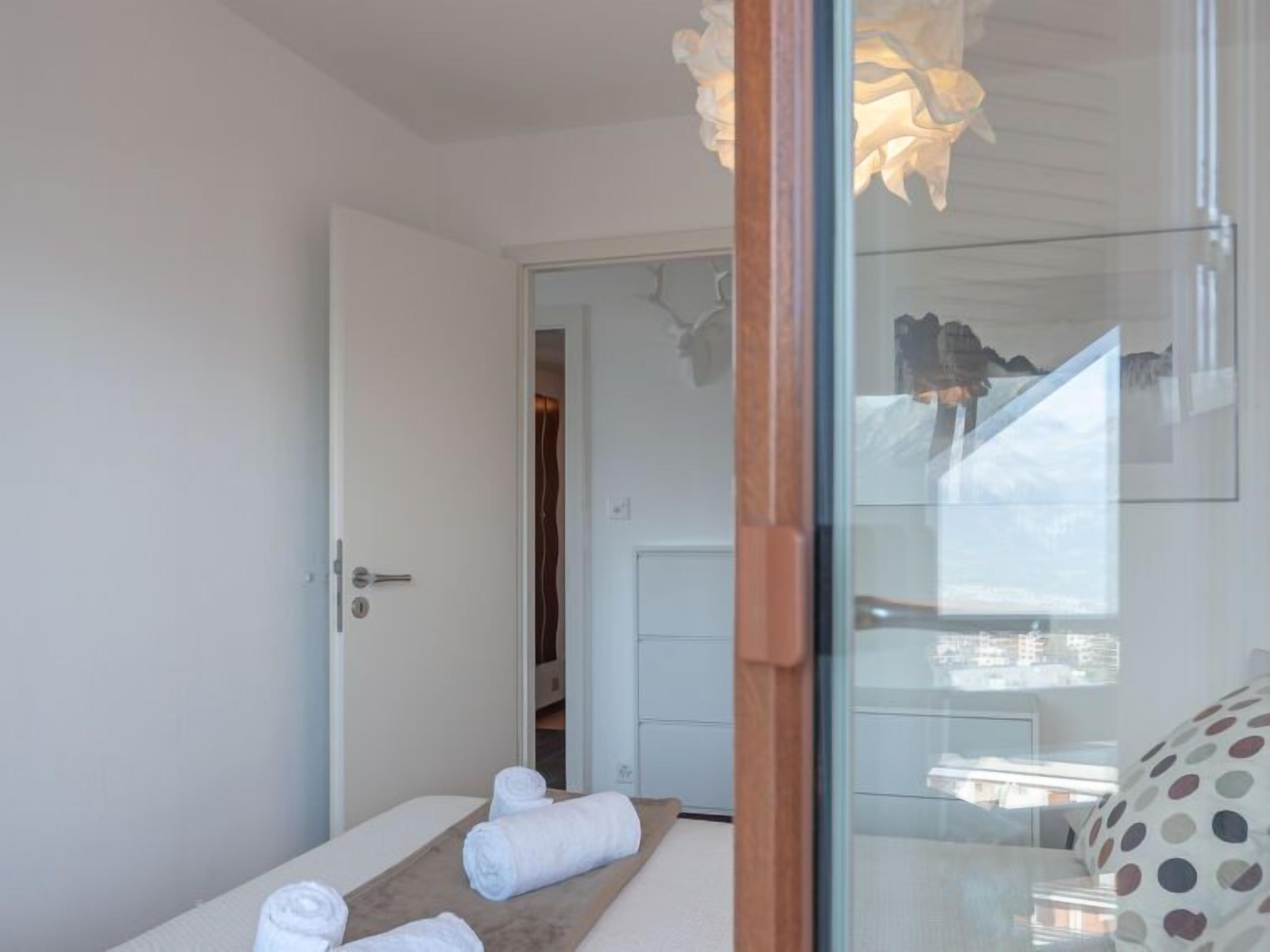 Une chambre avec un lit, une table de chevet en bois sur laquelle sont pliés des serviettes, une cloison en verre et une vue sur la ville par la fenêtre.