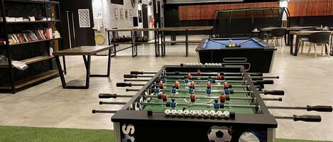 A room with a foosball table, a pool table, and a bookshelf with books.