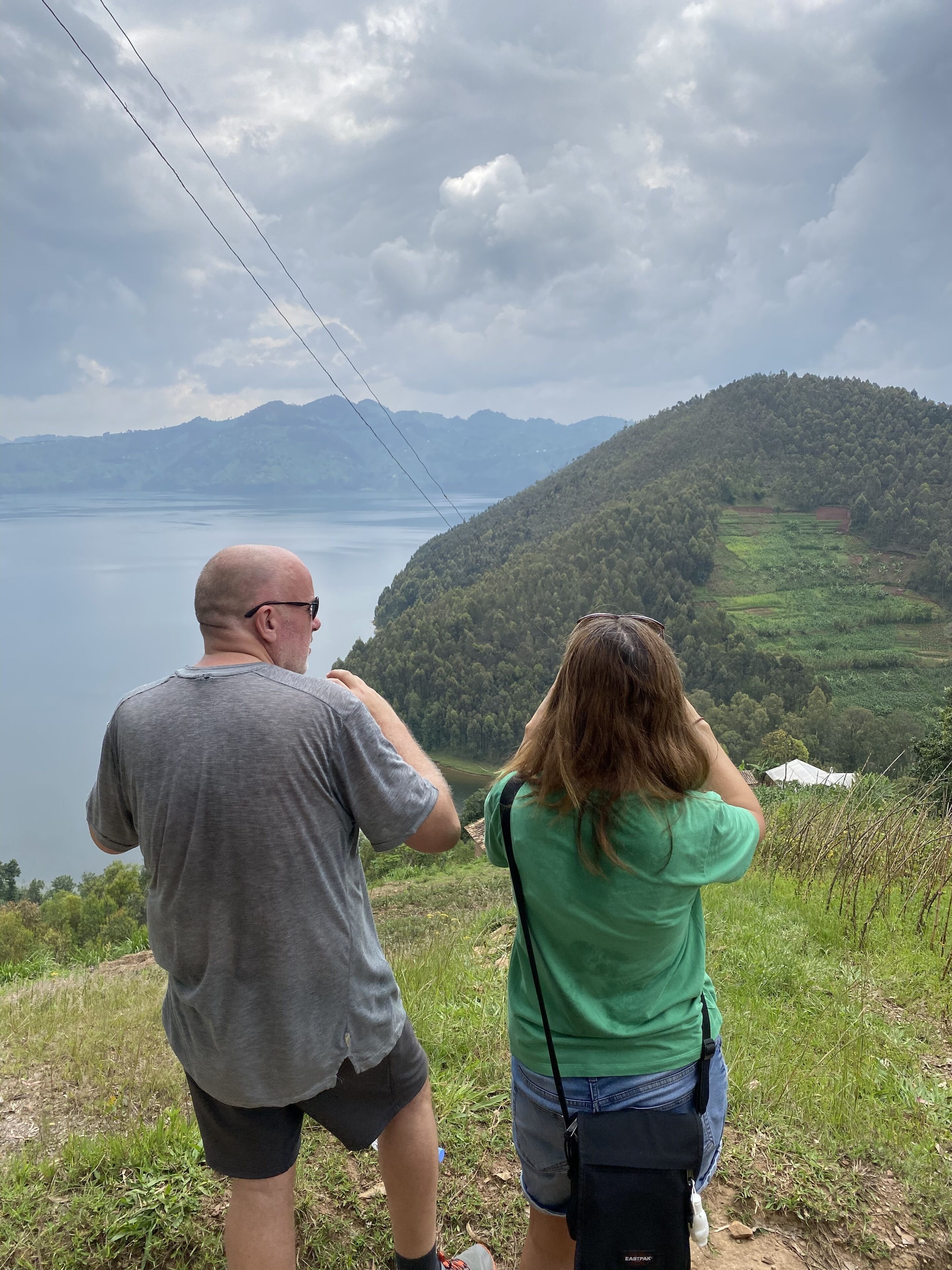 Two people standing on a grassy hill overlooking a lake and mountains.