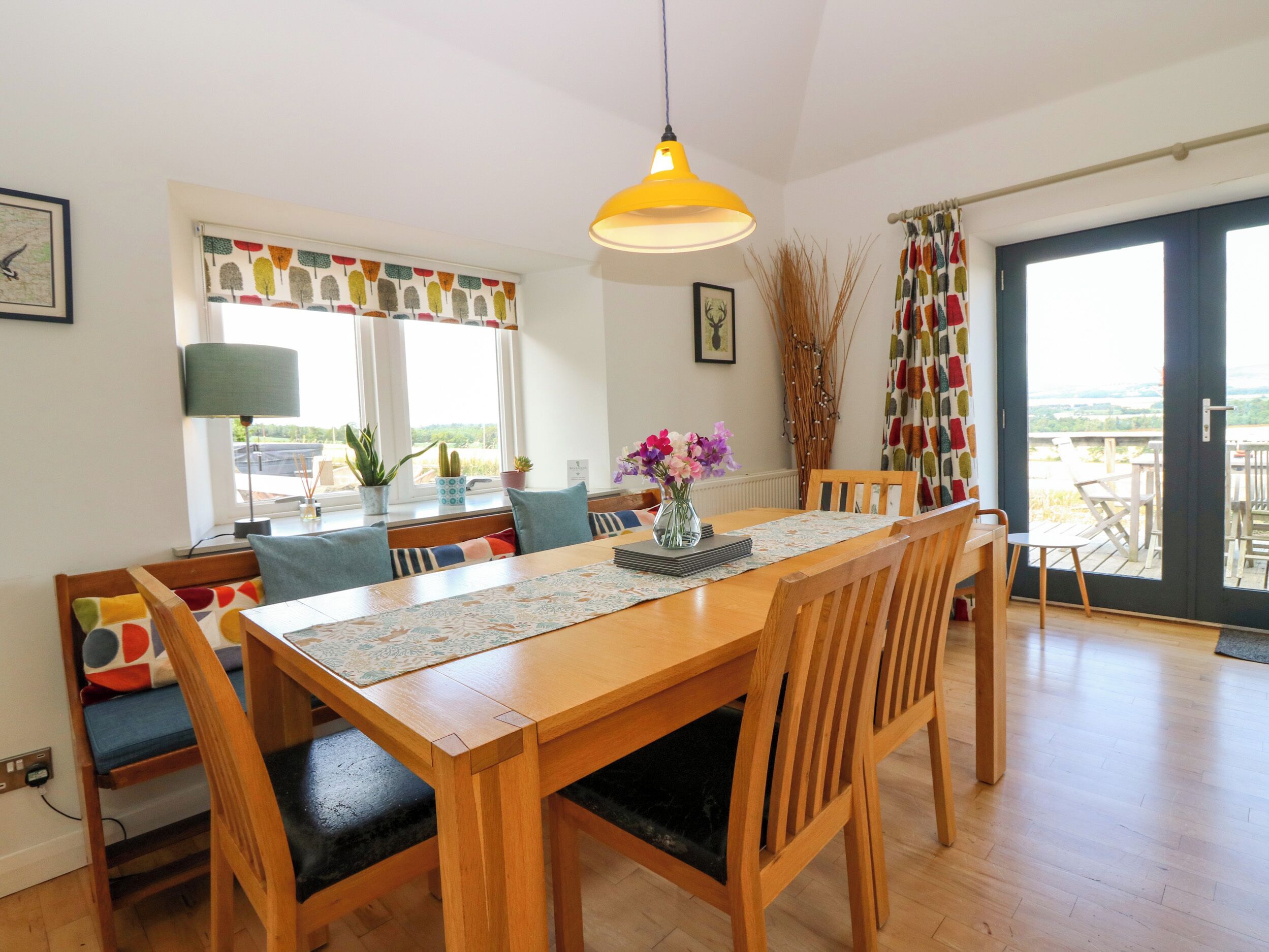 Un comedor con una mesa de madera, sillas, una lámpara colgante amarilla y vistas a través de un ventanal.