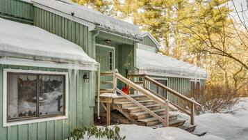 A green wooden cabin with a wooden staircase leading to a door, surrounded by snow and trees.
