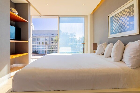 Quarto moderno com uma cama grande, uma televisão em uma estante e uma varanda com vista para a cidade.
