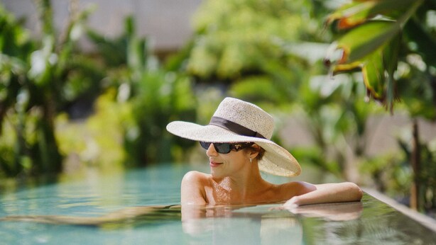 A person in a swimming pool with a hat, surrounded by greenery.