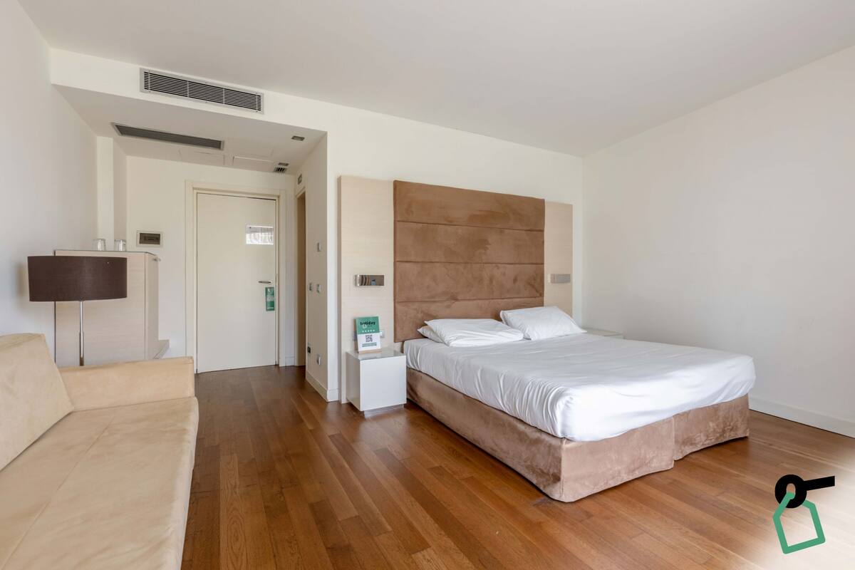 A bedroom with a large bed, a nightstand, a bedside table, a lamp, and a door leading to another room.