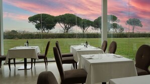 Un espace de restauration intérieur avec des tables disposées pour deux personnes, de grandes fenêtres offrant une vue sur un champ verdoyant et des arbres, et un plafond agrémenté d’une lucarne.