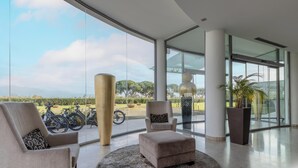 Une pièce spacieuse avec de grandes fenêtres, un coin salon avec des fauteuils et un pouf, et une vue sur des vélos et la verdure à l’extérieur.