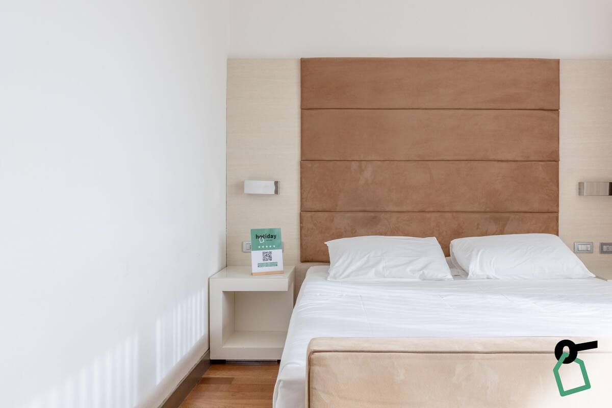 A hotel room with a large bed, a nightstand, a headboard, and a small table with a sign.