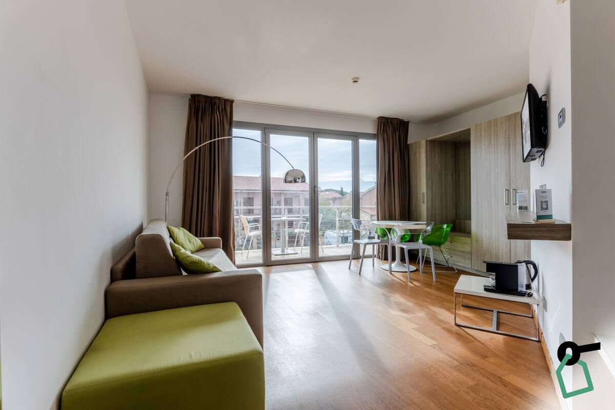A modern living room with a sofa, a dining table with chairs, a TV mounted on the wall, and a balcony with a view of buildings.