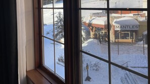 A window with a view of a snowy landscape, a building with a sign reading "MANTLES", and a clear blue sky. - Hearthstone Lodge #406 | 3 Peaks Rentals (Sun Peaks)