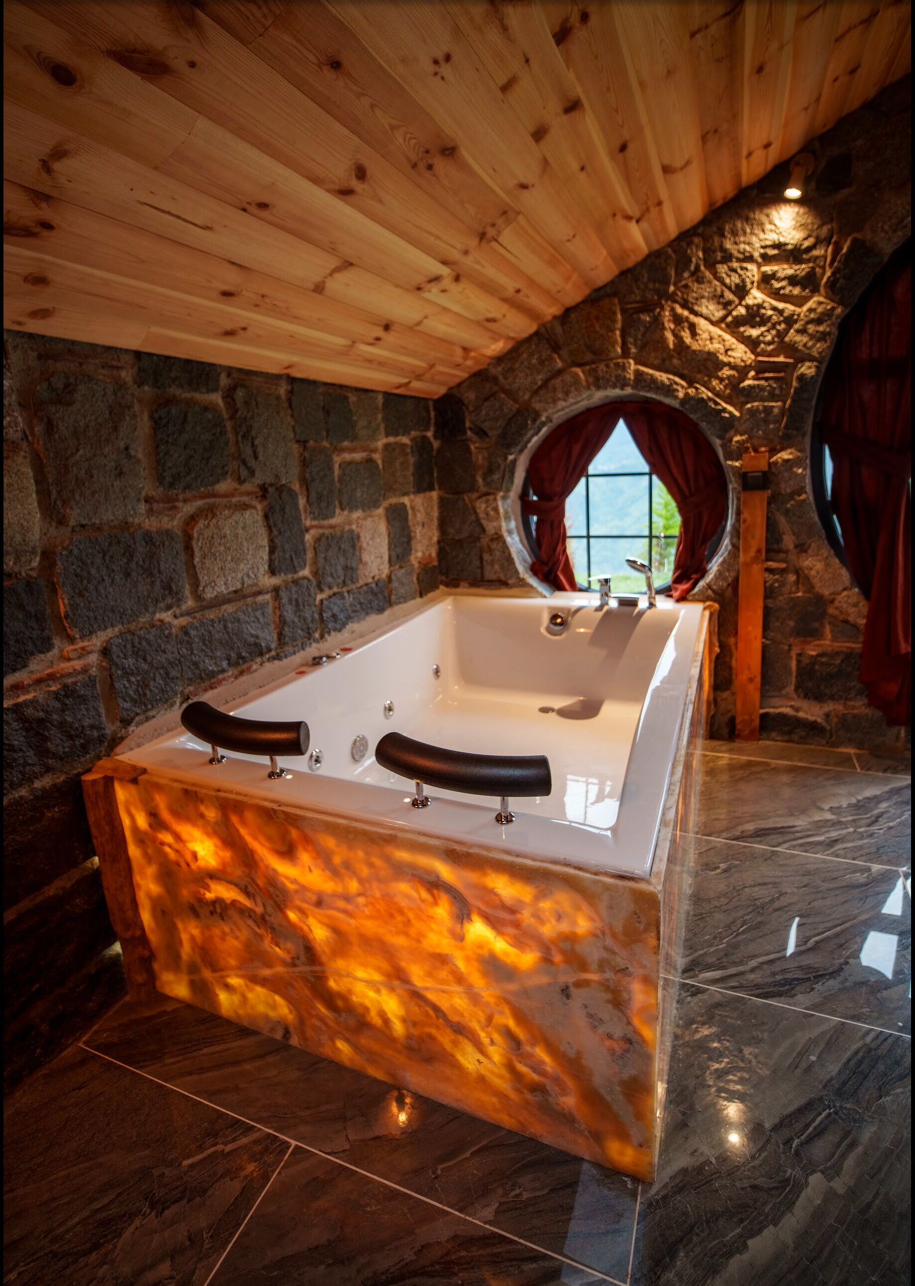 A stone-walled bathroom with a freestanding bathtub, a wooden ceiling, and a stone floor.