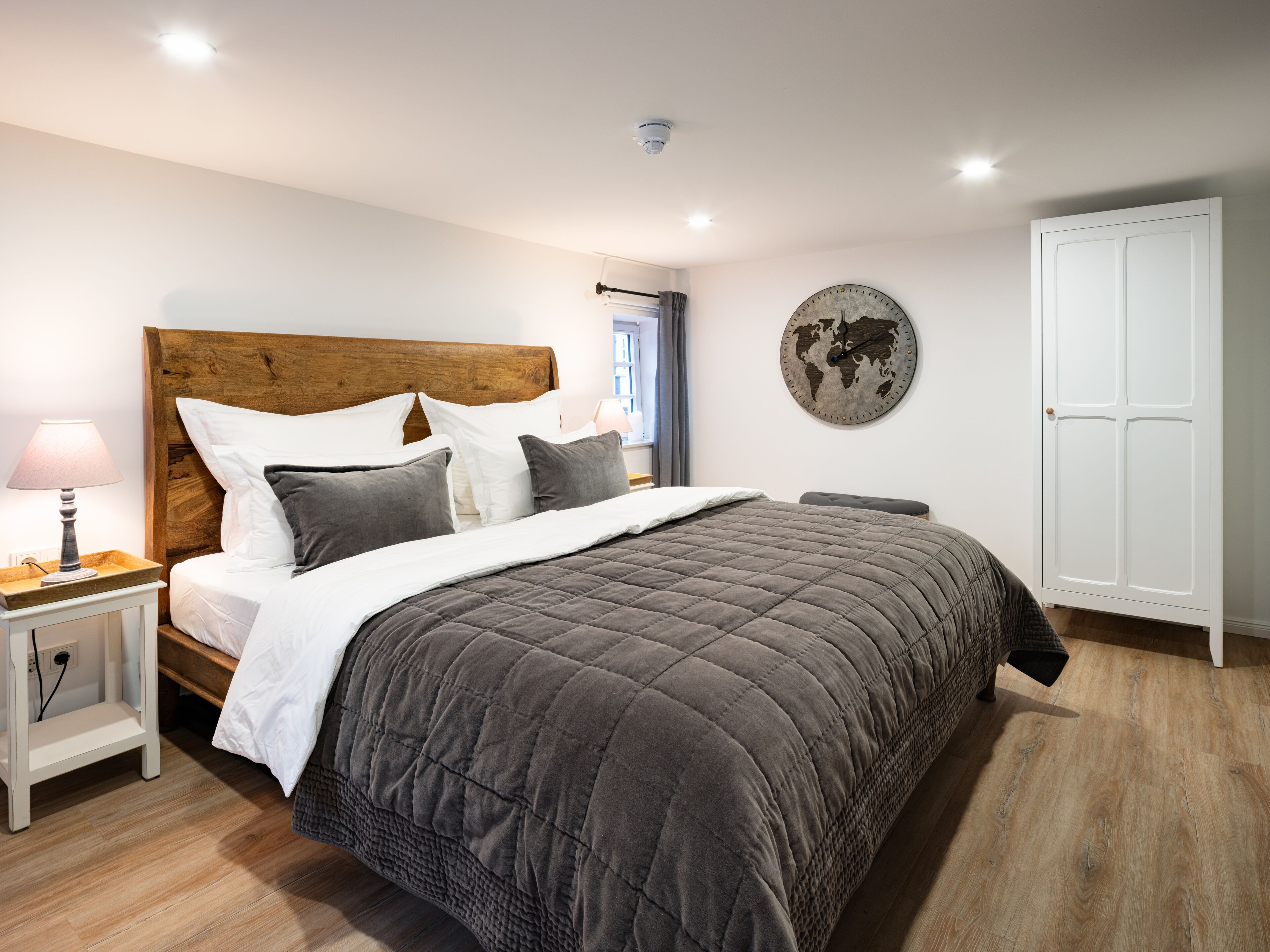 A bedroom with a wooden headboard, a large bed with a grey quilt, a bedside table with a lamp, a world map on the wall, and a white wardrobe.