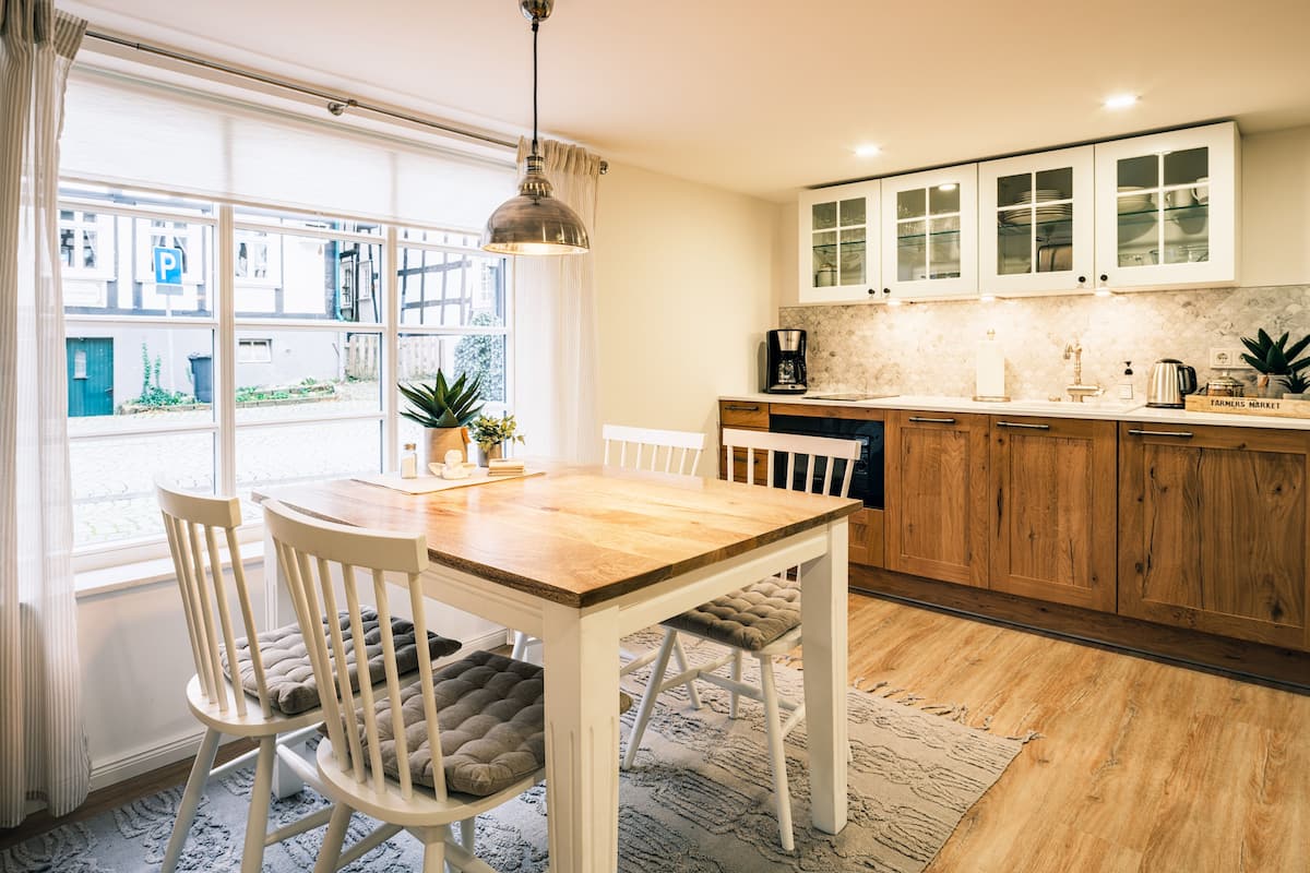 A modern kitchen with wooden cabinets, a dining table, and chairs. There is a window with a view of the street, and a plant on the windowsill.