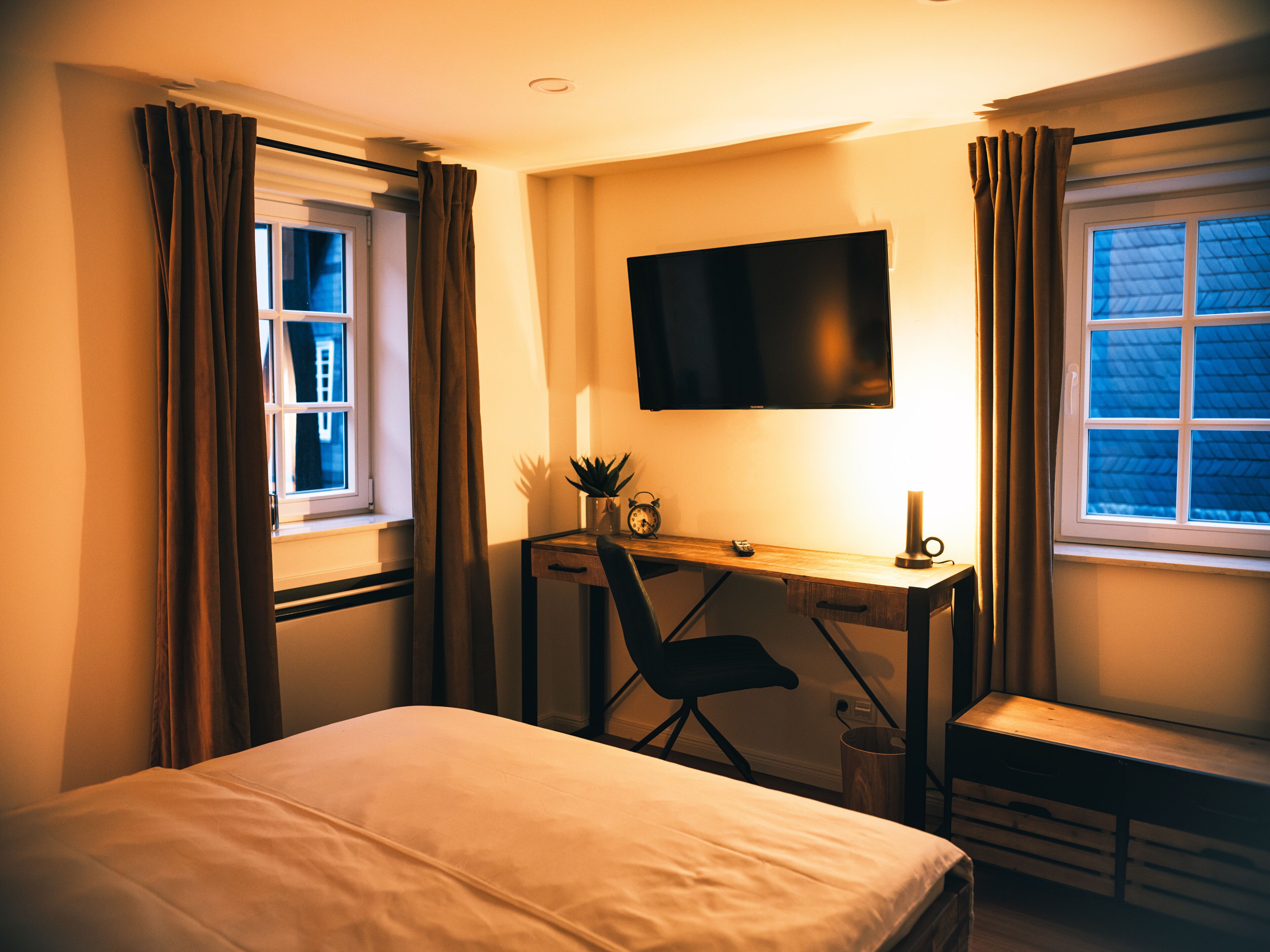 A hotel room with a bed, a desk with a chair, a TV mounted on the wall, and a window with curtains.