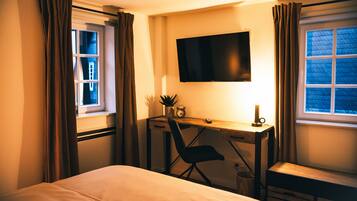 A hotel room with a bed, a desk with a chair, a TV mounted on the wall, and a window with curtains.