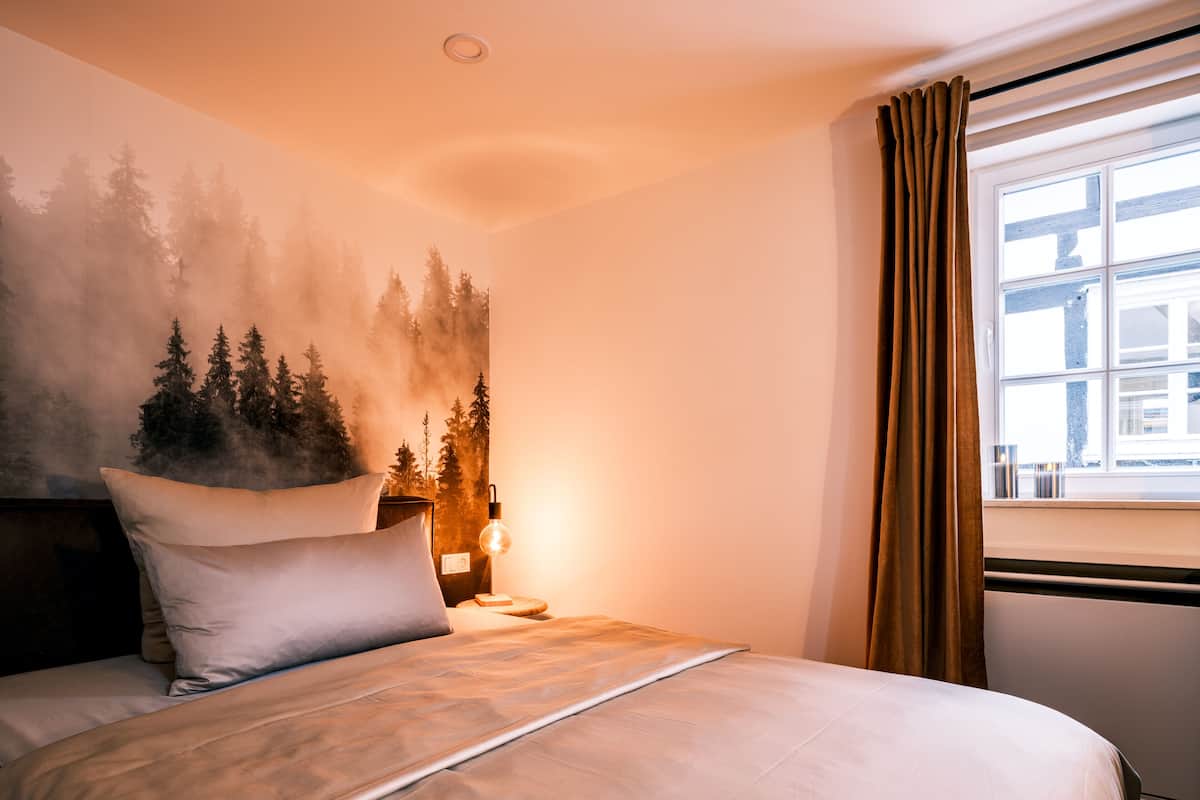 A neatly made bed with a headboard, a wall-mounted lamp, and a window with curtains.