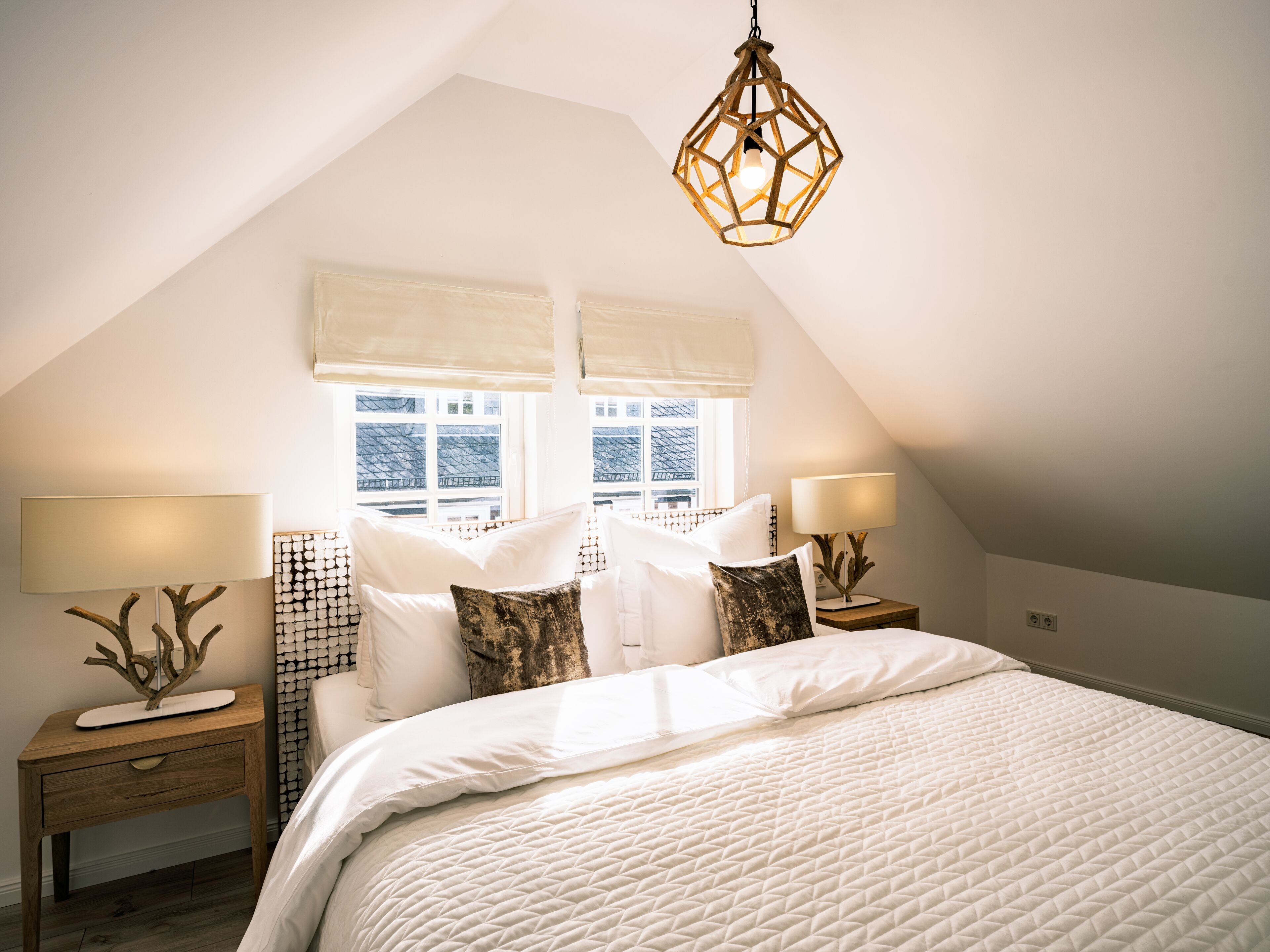 Une chambre à coucher avec un grand lit, deux tables de chevet, un luminaire suspendu et une fenêtre donnant sur des immeubles.