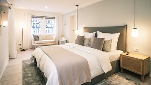 A modern bedroom with a large bed, bedside tables, a nightstand, a floor lamp, and a window with a view of buildings. - Flecker Hotel (Freudenberg)