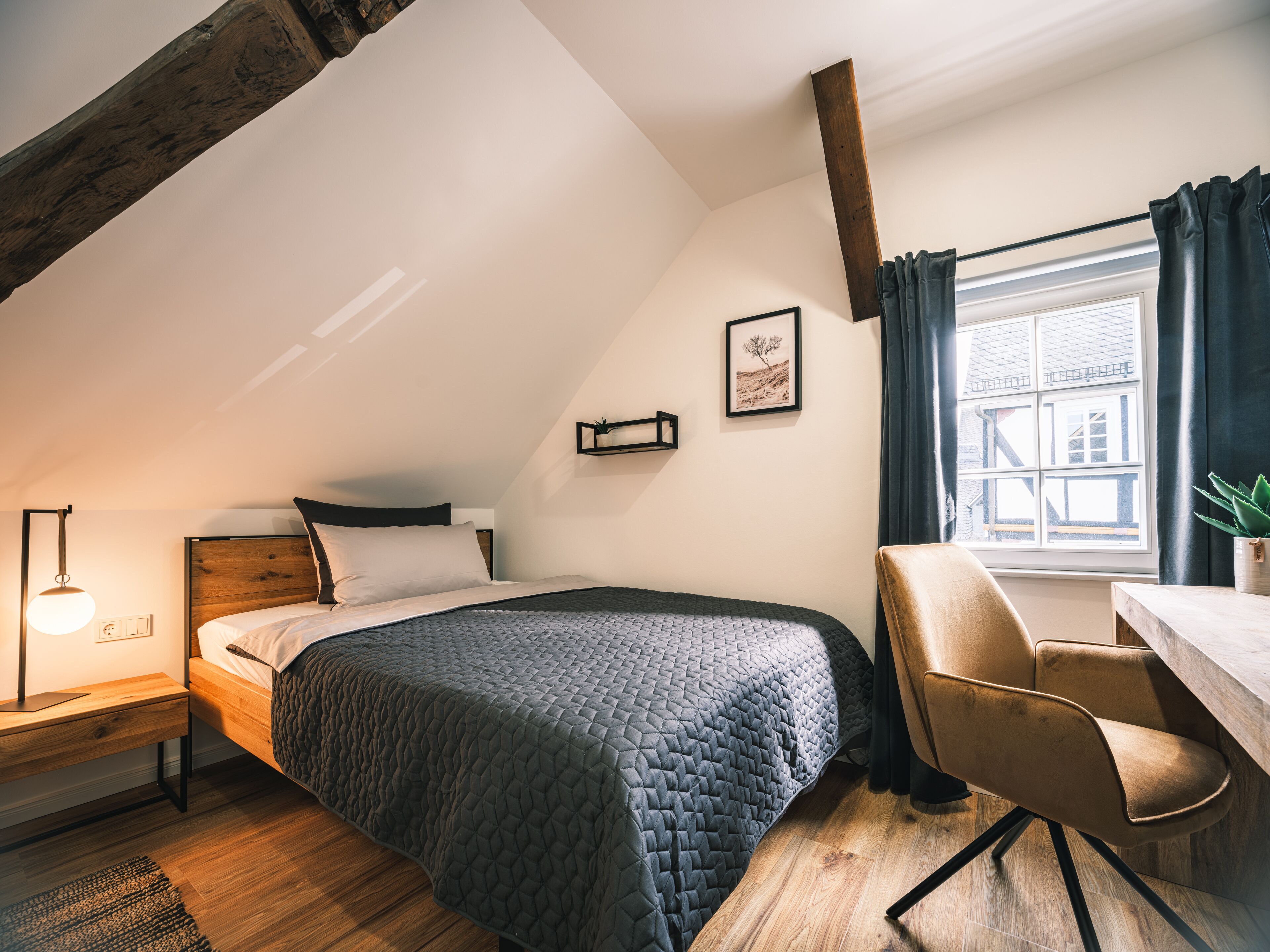 Een slaapkamer met een bed, een bureau, een stoel, een lamp en een raam met gordijnen.