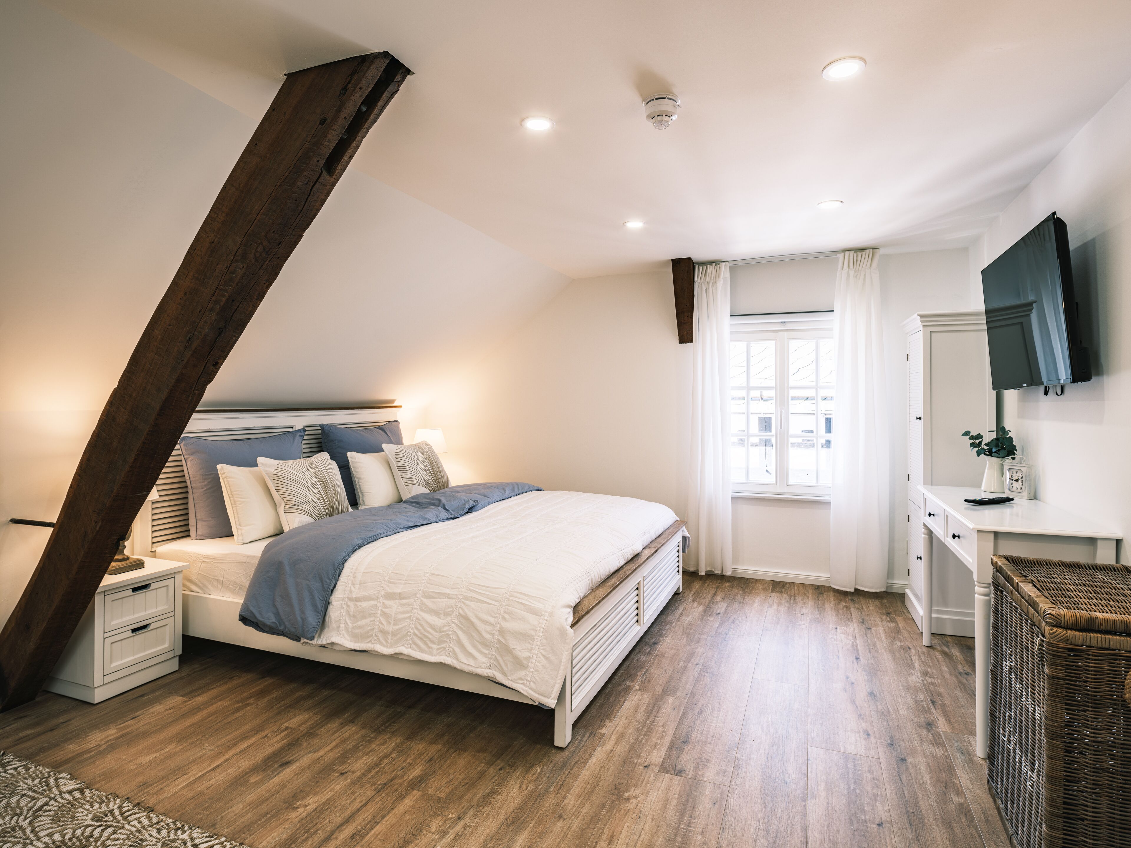 Ein Schlafzimmer mit schrägemaßtem Dach, einem Bett mit weißblauer Bettdecke, einem Nachttisch mit Lampe, einer Kommode, einem Korbwannen-Korb und einem an der Wand montierten Fernseher.
