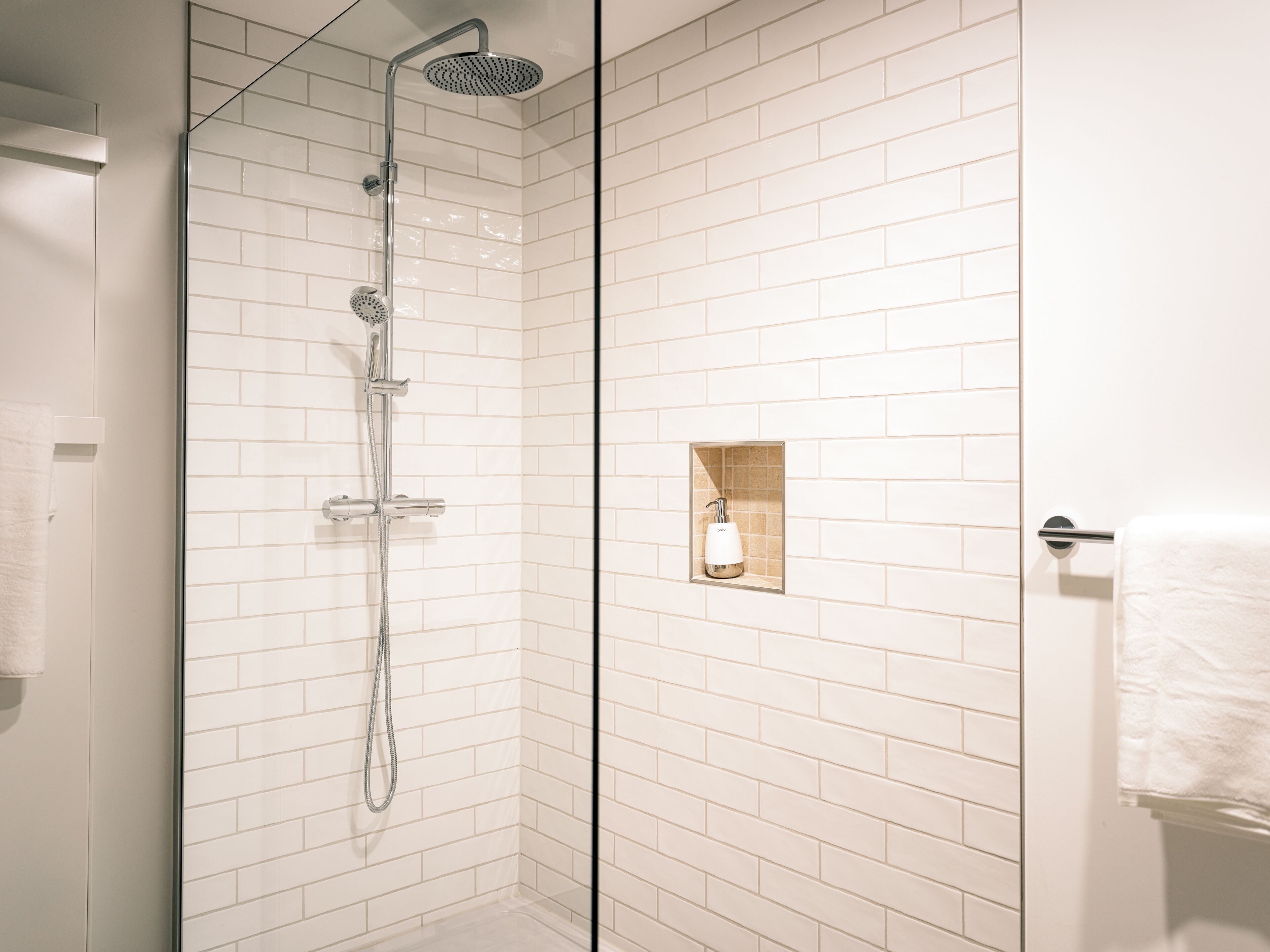 A modern shower with a glass door, white tiled walls, and a small shelf with a bottle.