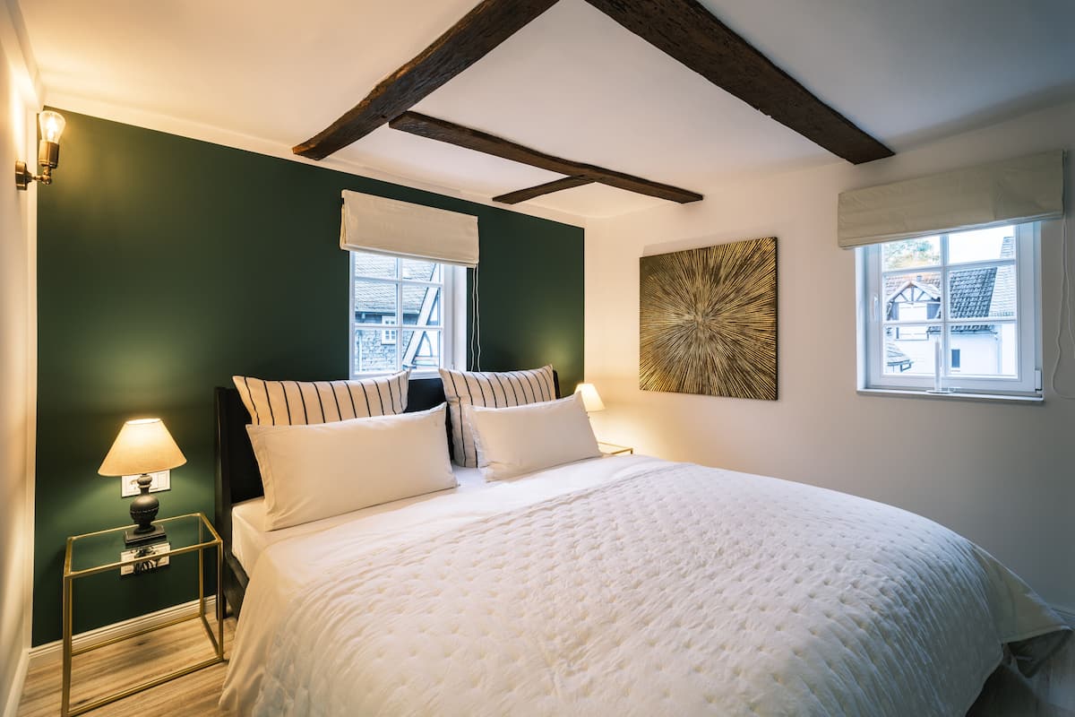 A bedroom with a bed, a nightstand, a lamp, a window with a view, and a wall with a geometric pattern.