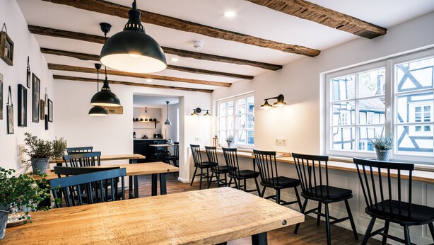 A dining area with wooden tables and black chairs, a kitchen in the background, and large windows with a view of buildings. - Flecker Hotel (Freudenberg)