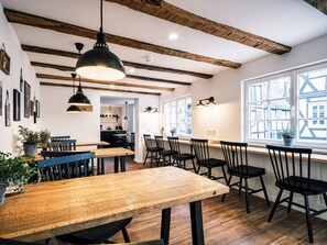 A dining area with wooden tables and black chairs, a kitchen in the background, and large windows with a view of buildings. - Flecker Hotel (Freudenberg)