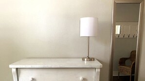 Een witte nachtkastje met een lamp, een rieten stoel en een kapstok op de achtergrond.
