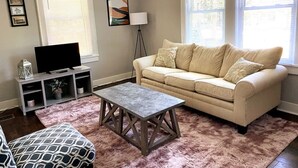 Een woonkamer met een beige bank, een salontafel, een televisie op een dressoir en een vloerlamp.