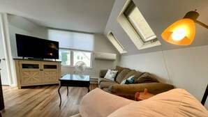 Una sala moderna con televisor de pantalla plana, sofá, mesa de centro y ventilador de techo. Hay tragaluces y una ventana que aportan luz natural.