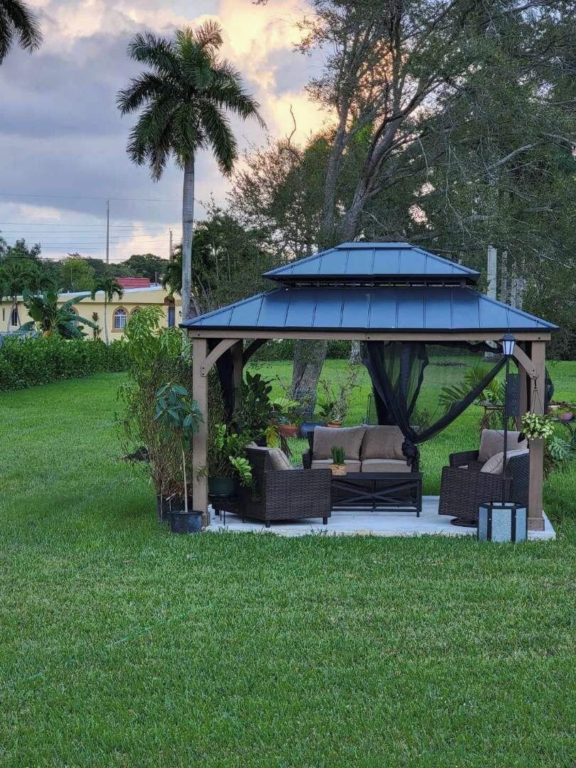 A gazebo with a roof, surrounded by a grassy area, and furnished with a sofa and chairs.