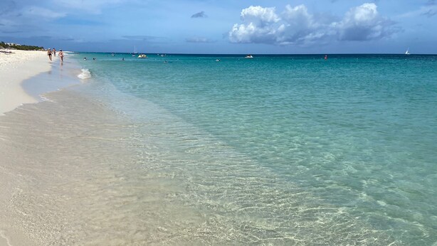 Une plage à l’eau turquoise et limpide, un rivage sablonneux et quelques bateaux aperçus au loin.