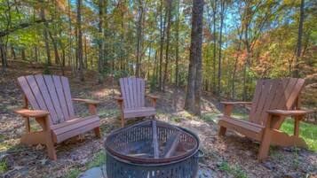 Three Adirondack chairs arranged around a fire pit in a forest setting.